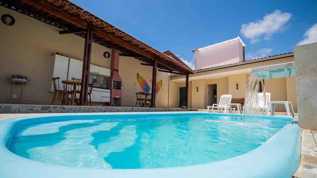 A clear blue swimming pool is framed by a stone patio, featuring a gentle waterfall feature. Sun loungers are positioned nearby, and an outdoor dining area can be seen in the background. The area is brightened by natural light and has a peaceful ambiance.