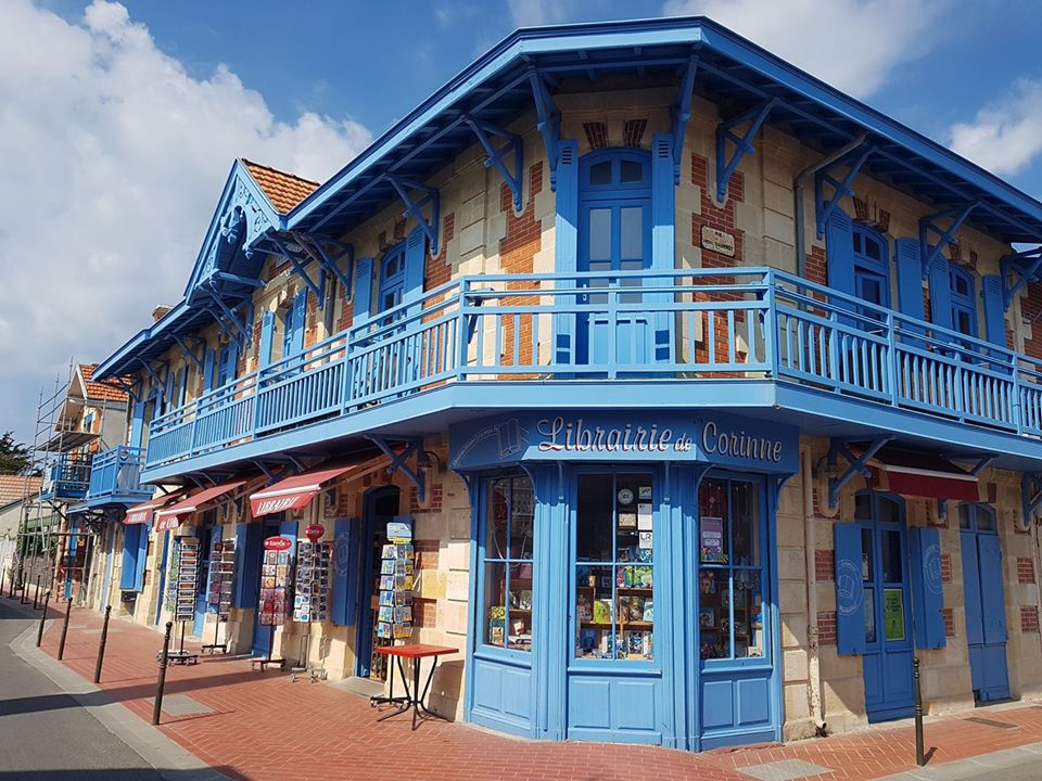 A charming blue building features a prominent bookstore on the ground floor. Its architectural details include decorative brickwork and spacious balconies. The surrounding area is pedestrian-friendly, lined with opportunities for shopping and exploring. A clear sky adds to the inviting atmosphere.