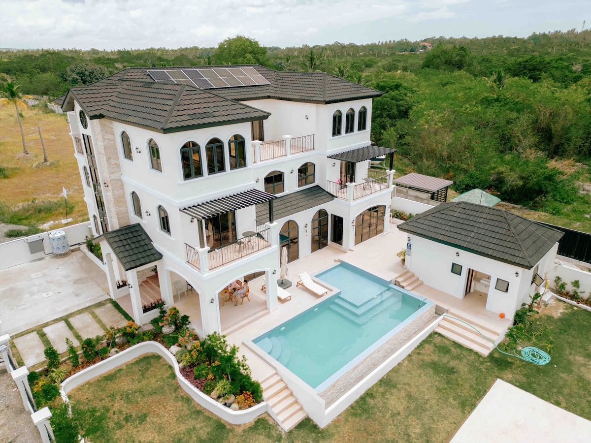 An aerial view showcases a multi-level home featuring a light exterior and a dark tiled roof. A swimming pool is positioned in the backyard, with a landscaped area surrounding it. Outdoor seating can be seen near the pool, offering an open and inviting space.