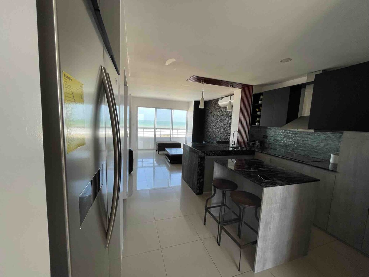 A modern kitchen features sleek cabinetry and a spacious island with bar stools. A large refrigerator is visible next to a window that allows natural light to illuminate the area. A view of the horizon can be seen through the expansive glass doors in the background.