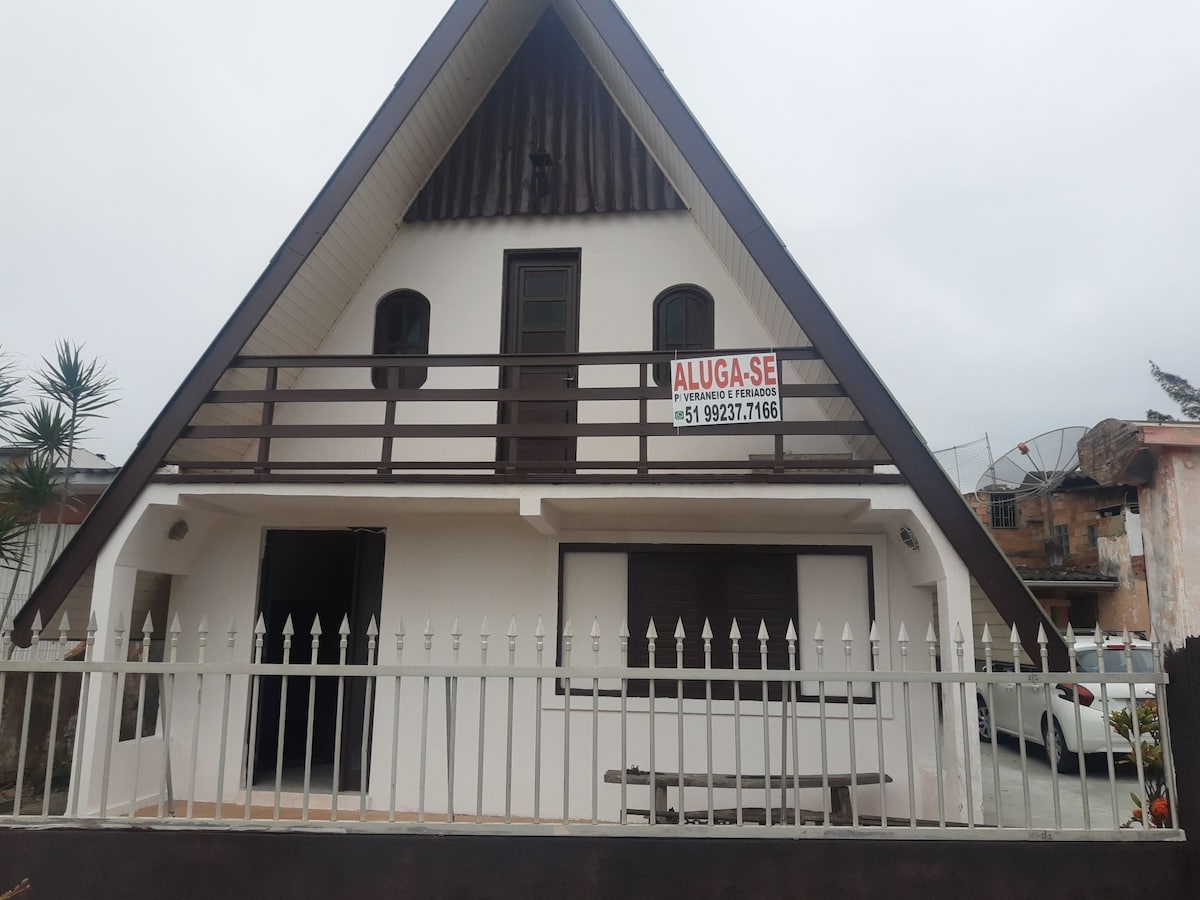 A triangular-roofed house features a spacious balcony with wooden railings. A 'For Rent' sign hangs prominently, while the exterior is painted in light colors. A small table is visible on a patio area, and the property is surrounded by a white picket fence, providing a sense of enclosure.