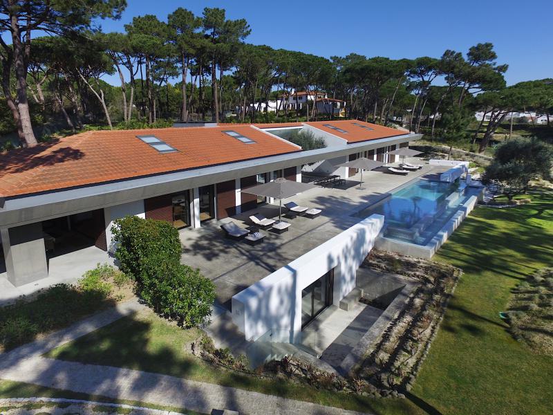A contemporary villa is set within a landscaped garden, featuring a modern design with a sloped red roof. An infinity pool extends alongside the spacious terrace, where sun loungers are arranged. Lush pine trees surround the property, offering a peaceful atmosphere.