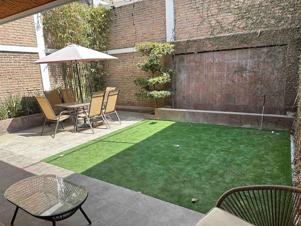 A serene outdoor space features a well-maintained artificial lawn and a dining area with a large umbrella. A round seating area is visible in the foreground, while lush greenery outlines the perimeter, providing a sense of privacy and tranquility.