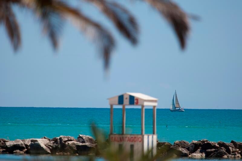 A distant sailboat is seen gliding across the calm turquoise waters. In the foreground, a lifeguard station with a colorful signage is positioned near the shoreline, framed by palm leaves swaying gently in the breeze.