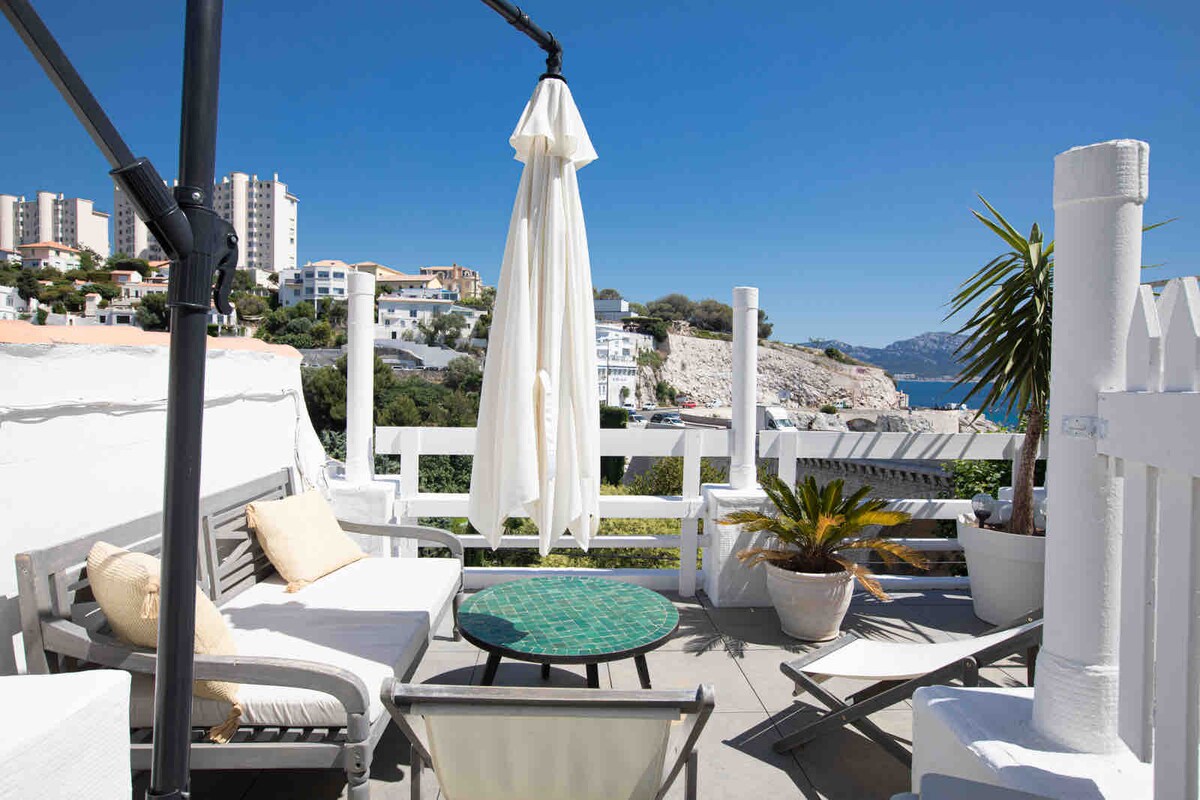 A private terrace is presented, featuring a round table with a mosaic top surrounded by comfortable seating. Potted plants add greenery, and a large umbrella provides shade. The backdrop includes a clear view of the sea and coastal landscape under a bright blue sky.