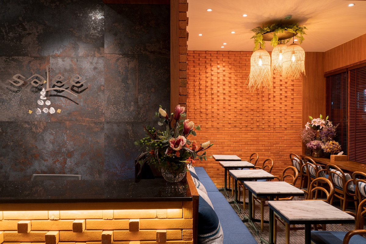 The reception area features a dark stone counter adorned with decorative plants and flowers. Behind it, a textured brick wall provides a warm background. Soft lighting is created by wicker pendant lamps, illuminating the space with a welcoming ambiance while tables are set for guests.