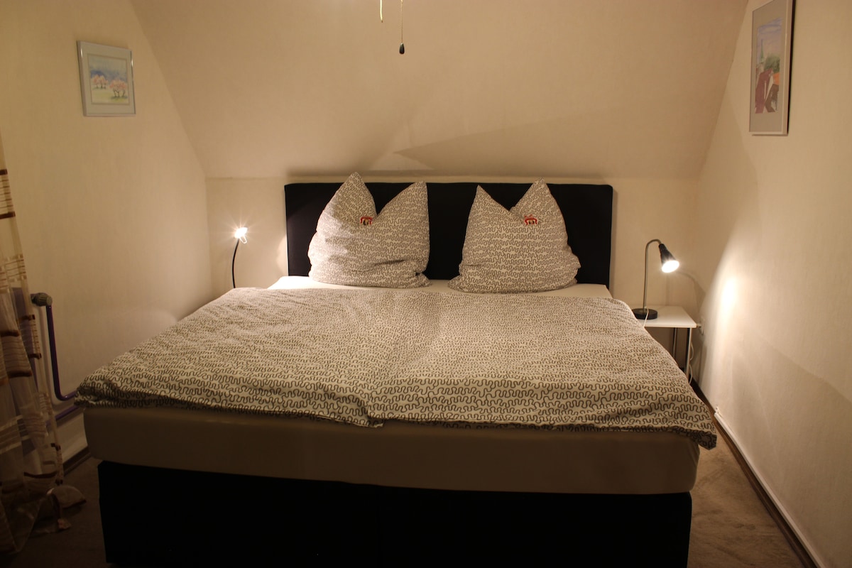 A comfortable sleeping area is presented with a spacious bed featuring two large, soft pillows. Two bedside lamps provide gentle illumination, and light-colored walls enhance the relaxing atmosphere. A small nightstand adds functionality to the space.