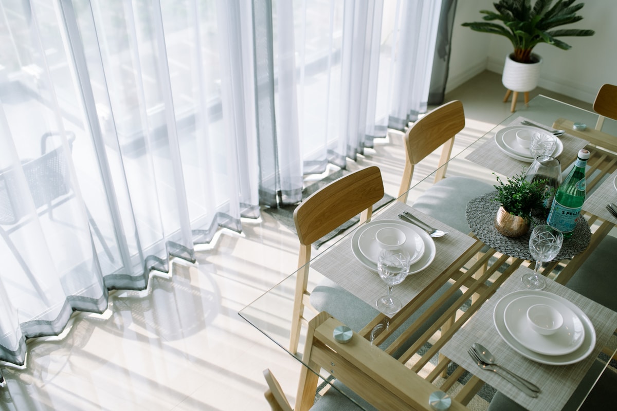 A dining area is presented with a glass table surrounded by wooden chairs. Elegant table settings include white plates and crystal glasses. Soft, sheer curtains allow natural light to filter through, while a small potted plant adds a touch of greenery.