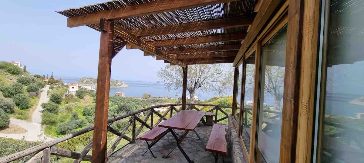 An outdoor terrace features a wooden roof and panoramic views of the sea and surrounding landscape. A long picnic table with wooden benches is positioned under the shelter, inviting relaxation while enjoying the natural scenery. The area is surrounded by greenery and traditional wooden railings.