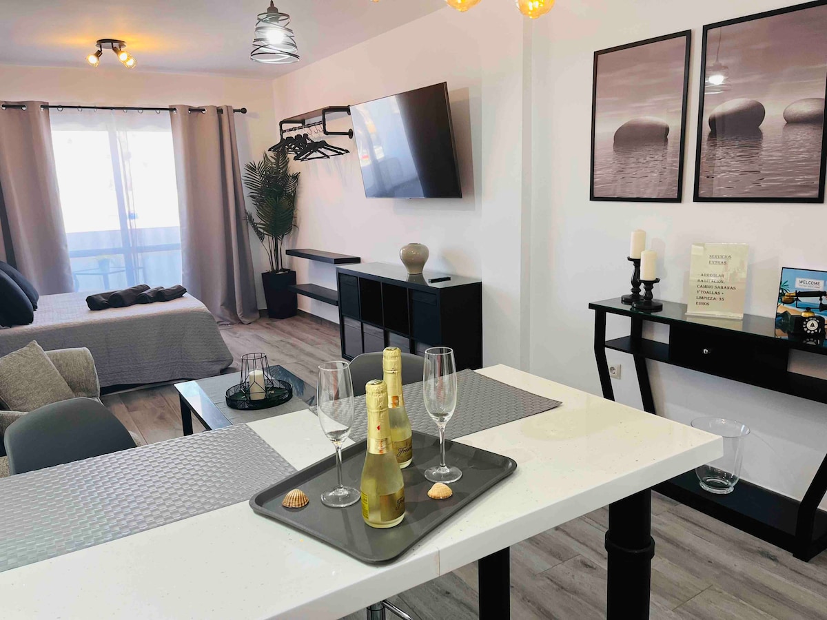 An inviting open-concept living area showcases a dining table with a decorative serving tray holding two flutes and a bottle of sparkling wine. A comfortable seating area is visible, along with a media unit and large windows allowing natural light to fill the space.