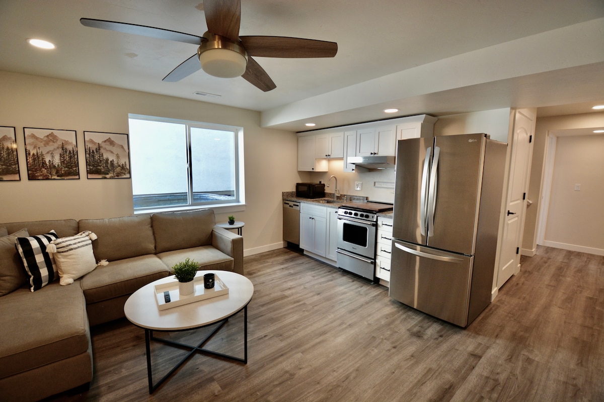 A modern kitchen is visually linked to a cozy living area, featuring a sectional sofa and a circular coffee table. A large window allows natural light to fill the space, while sleek stainless steel appliances and light cabinetry enhance the kitchen's functionality.
