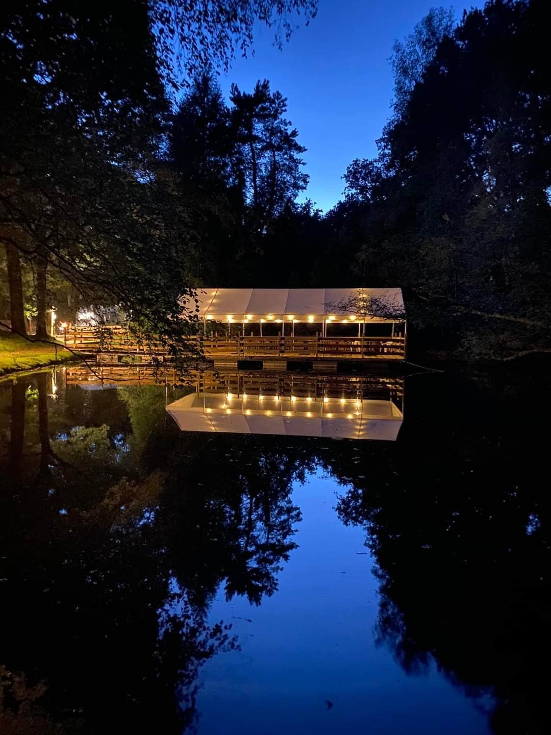 A charming structure is situated by a tranquil body of water, featuring soft lighting that reflects on the surface. Surrounding trees create a natural, serene backdrop, while the evening sky displays shades of blue, enhancing the peaceful ambiance.