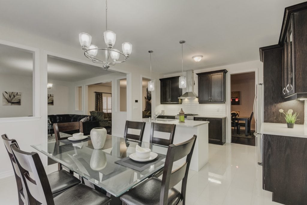 A modern kitchen and dining area is featured with a glass dining table set for six, surrounded by dark wooden chairs. Light fixtures hang above, complementing the cabinets and countertops. The space is open, with views into the adjacent living area, creating a seamless layout.