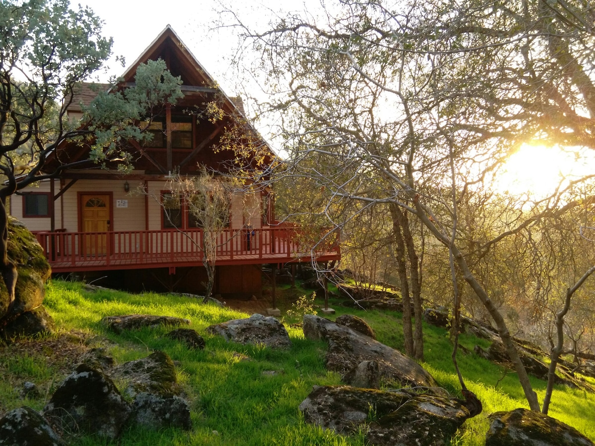 A charming cabin is nestled among lush green grass and large rocks. The structure features a sloped roof and a welcoming front porch with wooden railings. Soft sunlight filters through the trees, illuminating the scene with a warm glow.