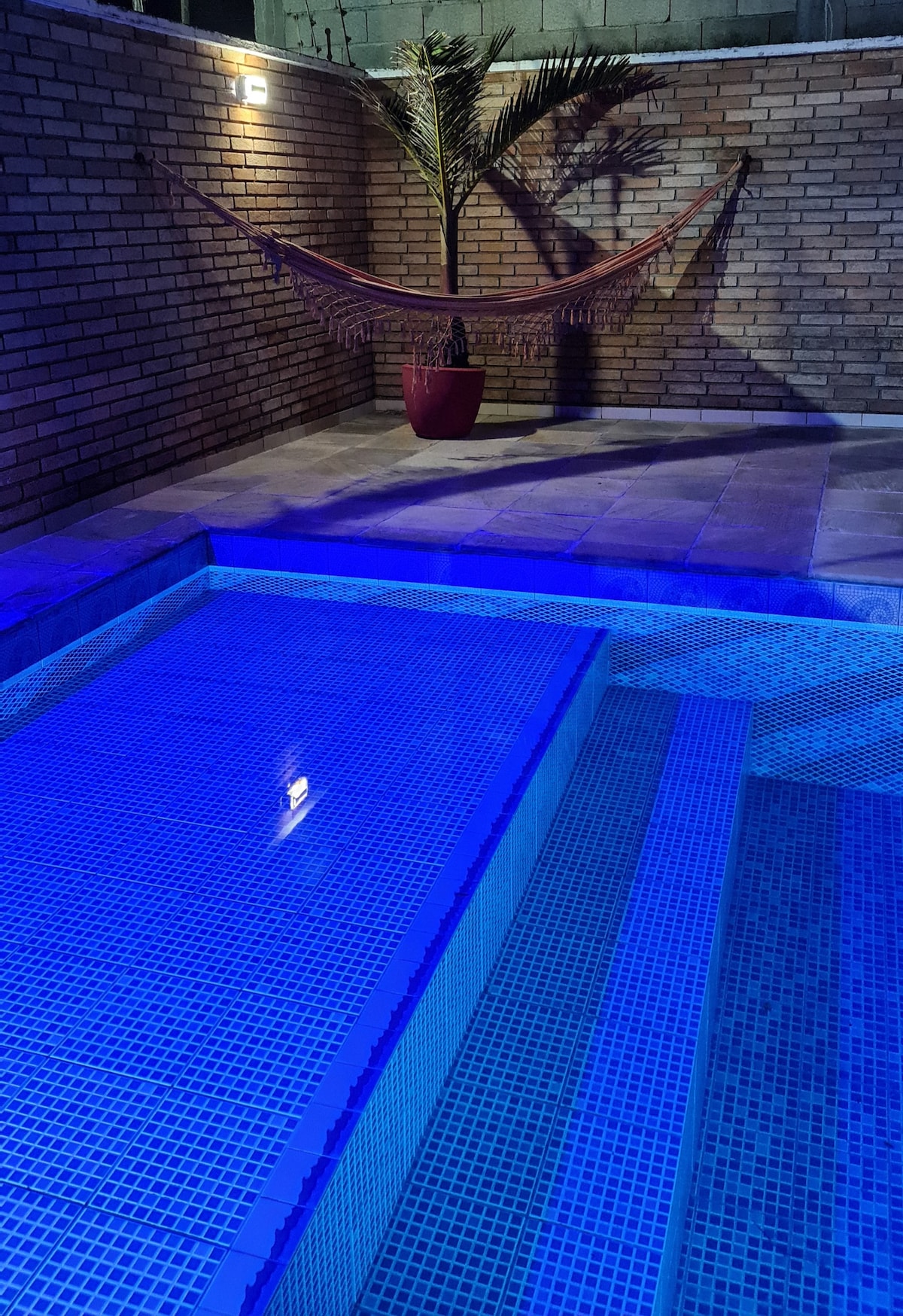 A tranquil pool area is illuminated with soft blue lighting, enhancing the inviting nature of the space. Steps lead into the water, while a hammock is positioned beside the pool, creating a relaxing atmosphere against a backdrop of textured brick walls.