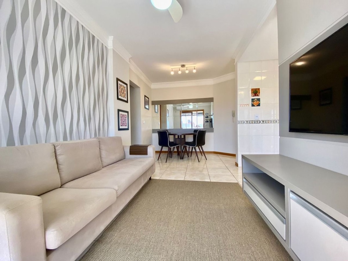A light-filled living area features a neutral-colored sofa and a flat-screen television. A dining table with six chairs is positioned adjacent to the sofa, with decorative wall art displayed on the walls. The mint-green accent wall adds a subtle touch of color.