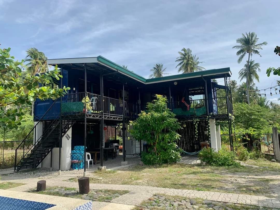 The exterior of the two-story container home is presented, surrounded by lush greenery and palm trees. A staircase leads to a balcony with seating. Casual outdoor furniture is visible on the ground level. The property showcases an open space with easy access to nature.