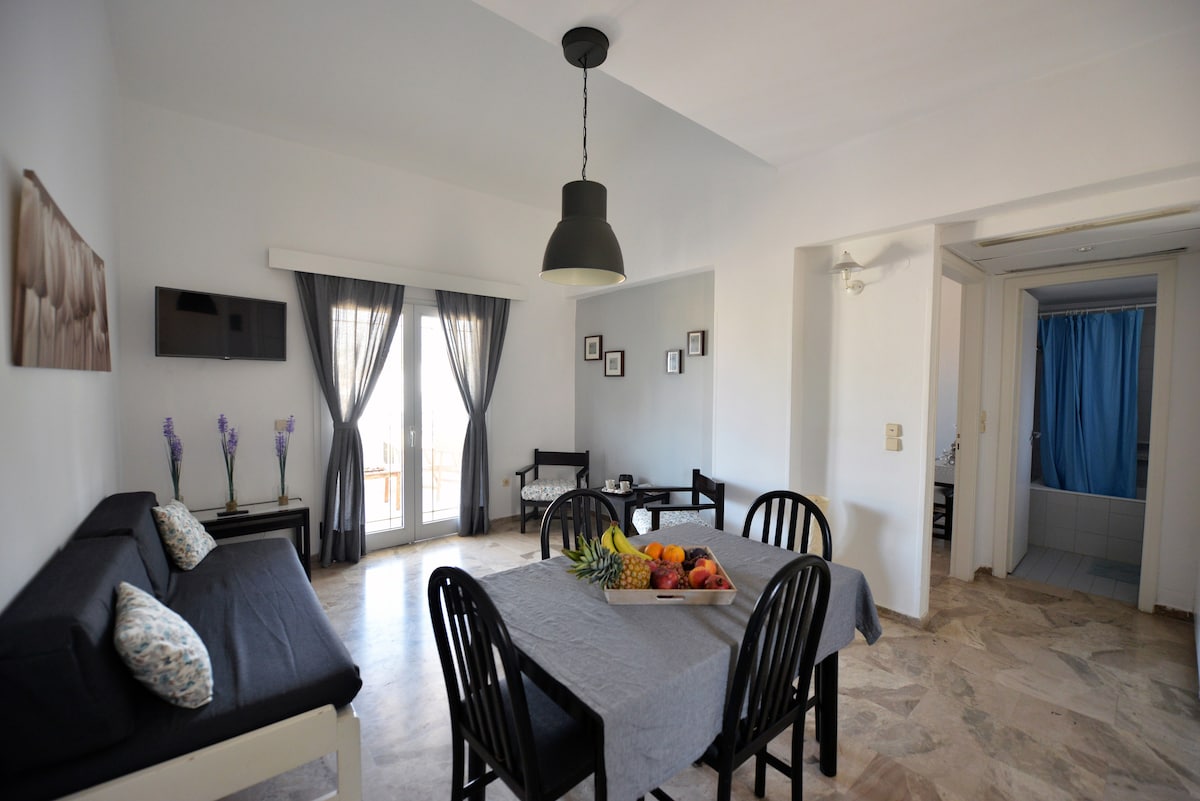 A dining area features a rectangular table surrounded by black chairs. A tray of fresh fruit is placed at the center. A cozy seating arrangement with a dark sofa is positioned nearby. Large glass doors provide access to a balcony, creating a light-filled environment.