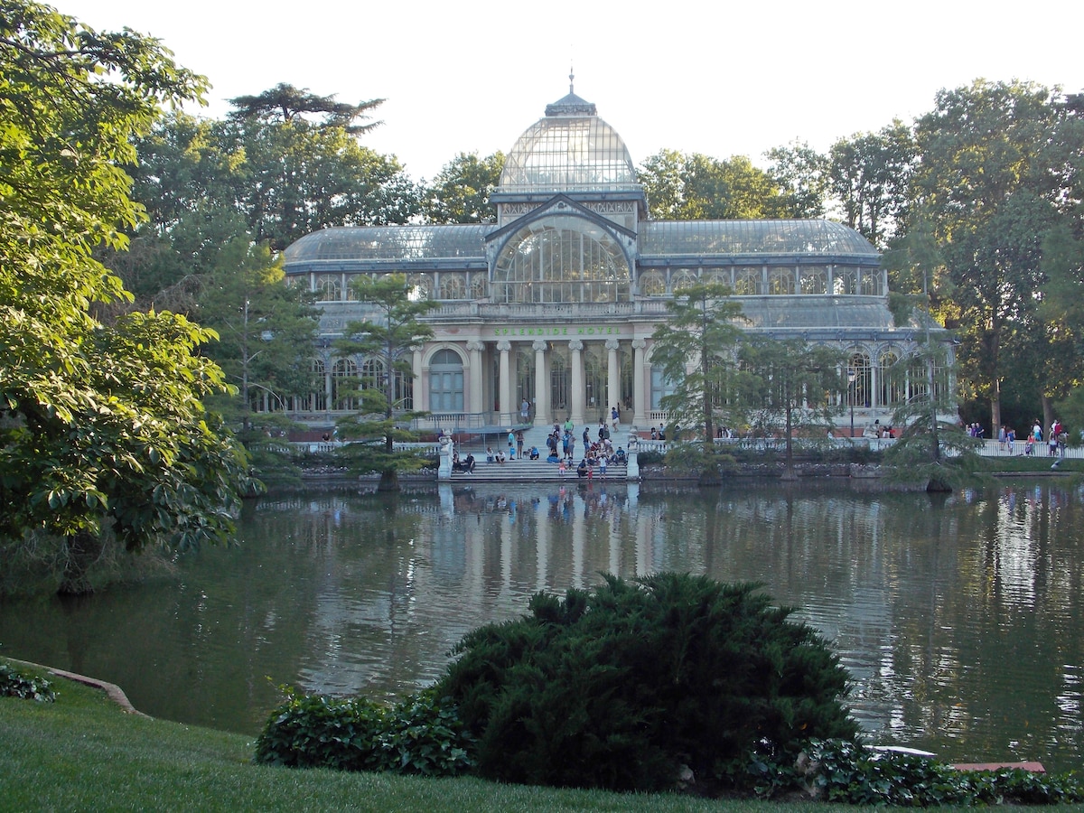 The image depicts a large glass structure surrounded by lush trees, reflecting on a tranquil lake. Visitors can be seen enjoying the park, with pathways leading towards the building, creating a serene atmosphere amidst greenery.
