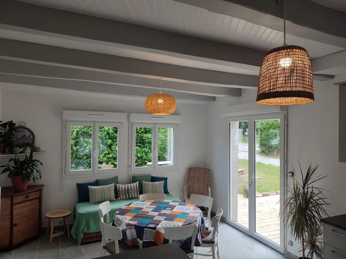 A communal living area features a large round table adorned with a colorful tablecloth. Light from multiple windows illuminates the space, revealing a cozy seating area with cushions. Two woven pendant lights hang from the ceiling, adding to the inviting ambiance.