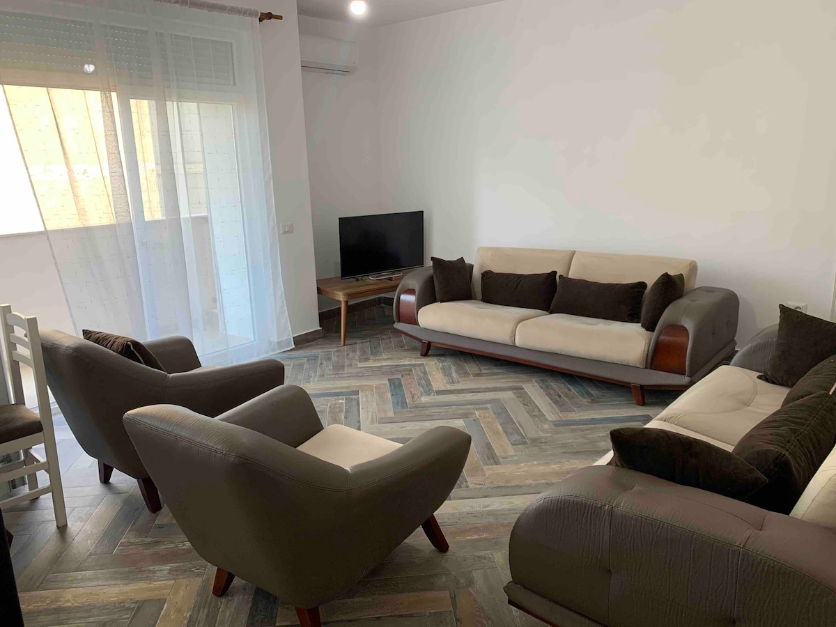 A cozy living area features two spacious sofas and two armchairs, all arranged around a wooden coffee table. A flat-screen TV is positioned on a small stand. Natural light is allowed in through large glass doors, with light-colored curtains softly filtering the sunlight.