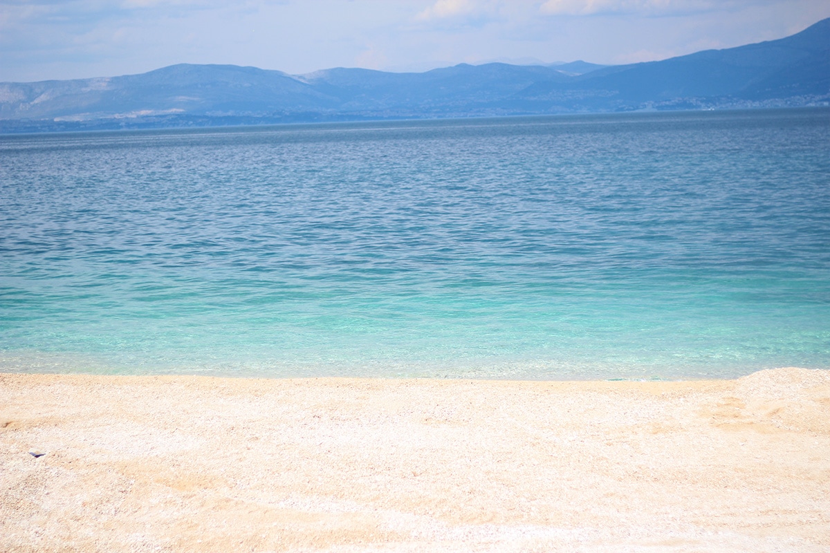 A serene shoreline is depicted, featuring soft sandy beach that transitions into clear turquoise water. The calm sea reflects the surrounding landscape, with distant mountains visible under a light blue sky, creating a tranquil setting ideal for relaxation.