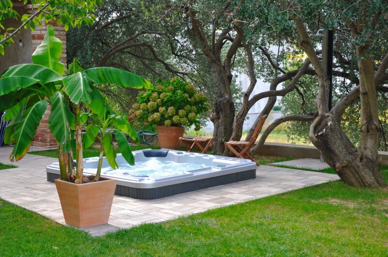 An outdoor area features a hot tub surrounded by greenery, including vibrant green banana plants and mature olive trees. A paved surface provides a comfortable setting, while two wooden loungers invite relaxation amidst the tranquil garden atmosphere.