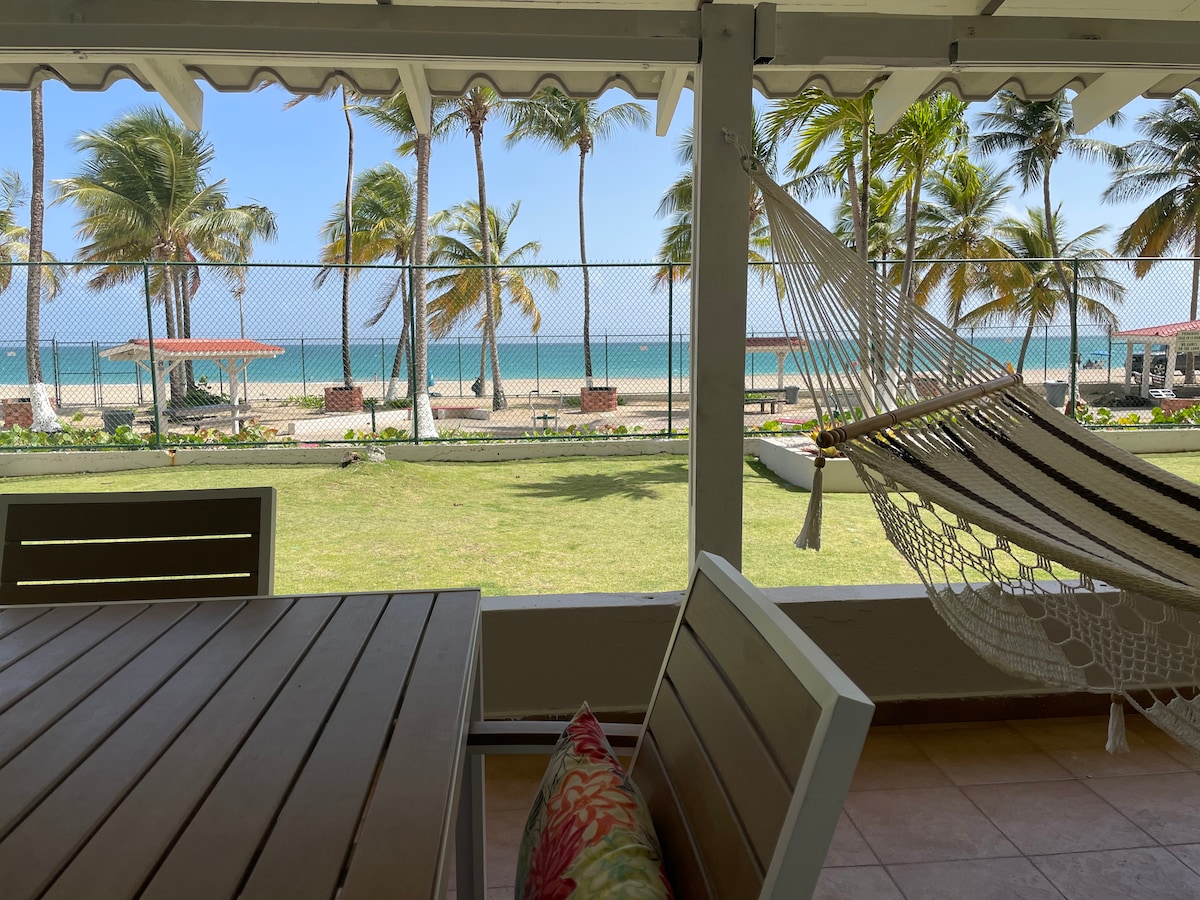 A serene outdoor seating area is seen through an open patio. A large hammock is suspended nearby, while palm trees sway gently in the breeze. In the background, the sparkling ocean is visible beyond a grassy area, creating a tranquil beachfront atmosphere.