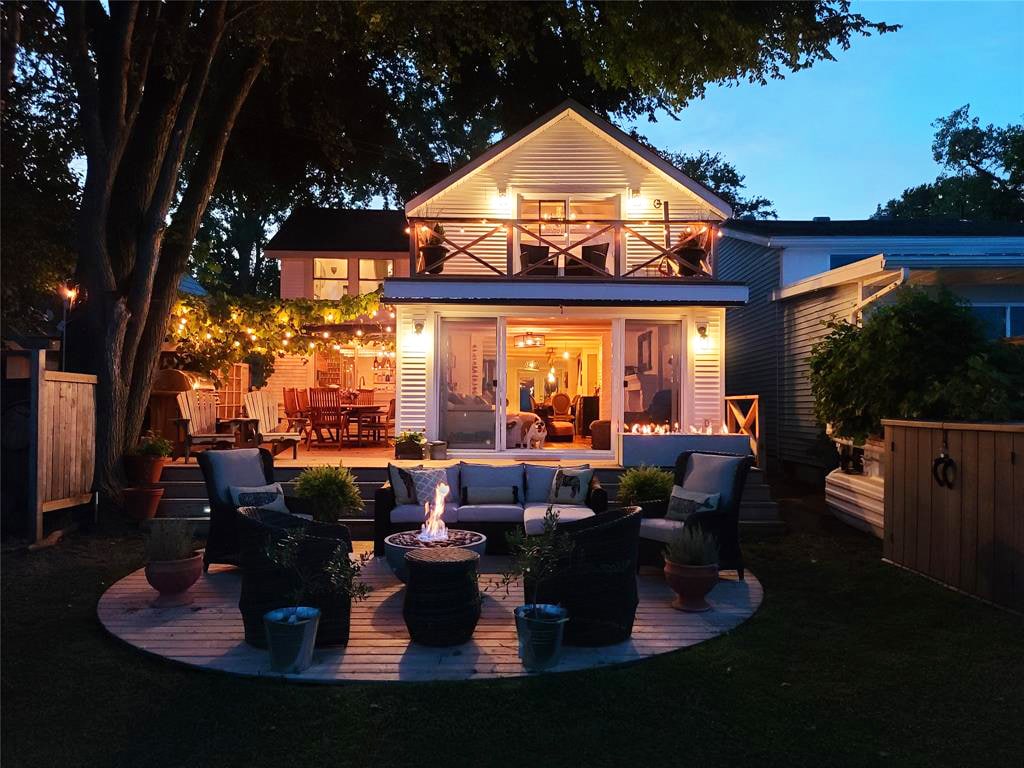 A charming outdoor space is illuminated by soft lighting from the house. A circular patio with wicker seating surrounds a fire pit, providing a welcoming area for relaxation. The house features large windows that reveal a cozy interior, with a balcony above offering additional seating.