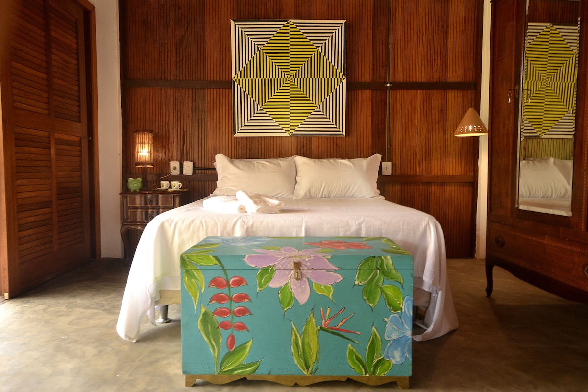 A cozy sleeping area features a spacious bed dressed in white linens, with colorful wall art above. A painted chest sits at the foot of the bed, showcasing vibrant floral designs. Natural wood paneling creates a warm atmosphere, complemented by a mirror and soft lighting.