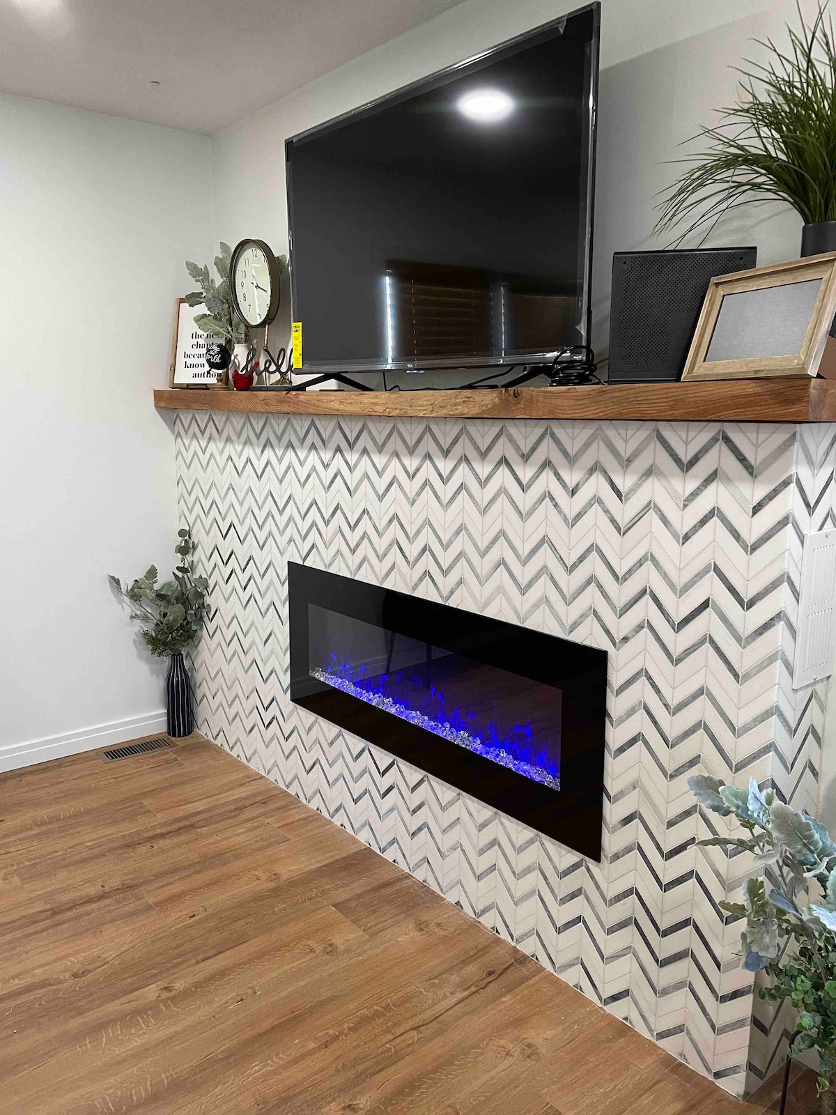 A stylish electric fireplace is displayed within a chevron-patterned wall, featuring blue LED accents. Above, a flat-screen television is positioned on a wooden shelf, accompanied by decorative elements such as a clock and potted plants. Natural light enhances the warm tones of the luxury vinyl flooring.