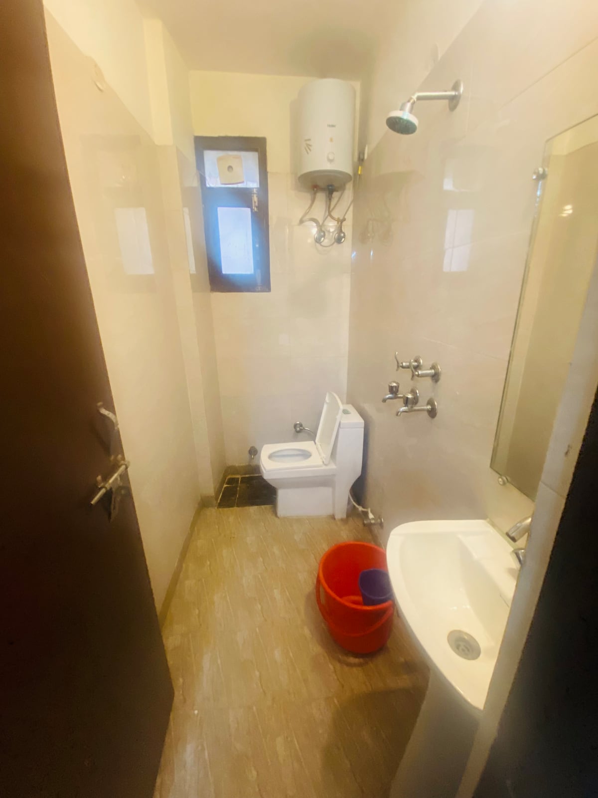 A compact bathroom is depicted, featuring a standard toilet and a sink with a faucet. A showerhead is installed above, and a mirror is mounted on the wall. A red bucket is shown on the floor, indicating the bathroom's utilitarian design.