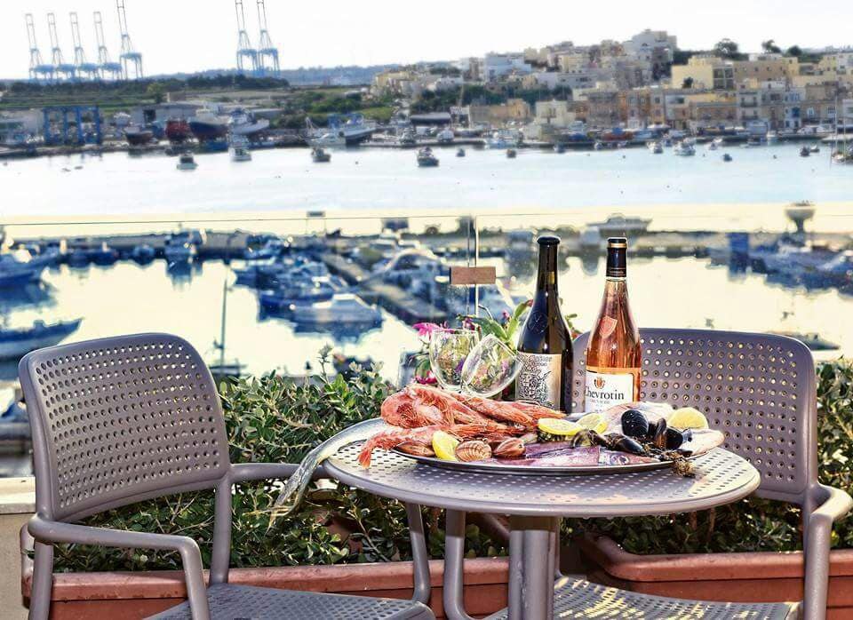 A table is set for dining, featuring seafood and beverages, with a picturesque view of Marsaxlokk Harbour and moored boats in the background. Two metal chairs accompany the table, surrounded by greenery. The scene captures a relaxing atmosphere in a coastal setting.