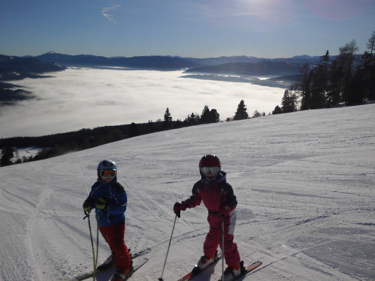 Two children are seen skiing on a snowy slope, wearing helmets and ski gear. A vast valley is visible in the background, covered in a layer of mist, with distant mountains under a clear blue sky.