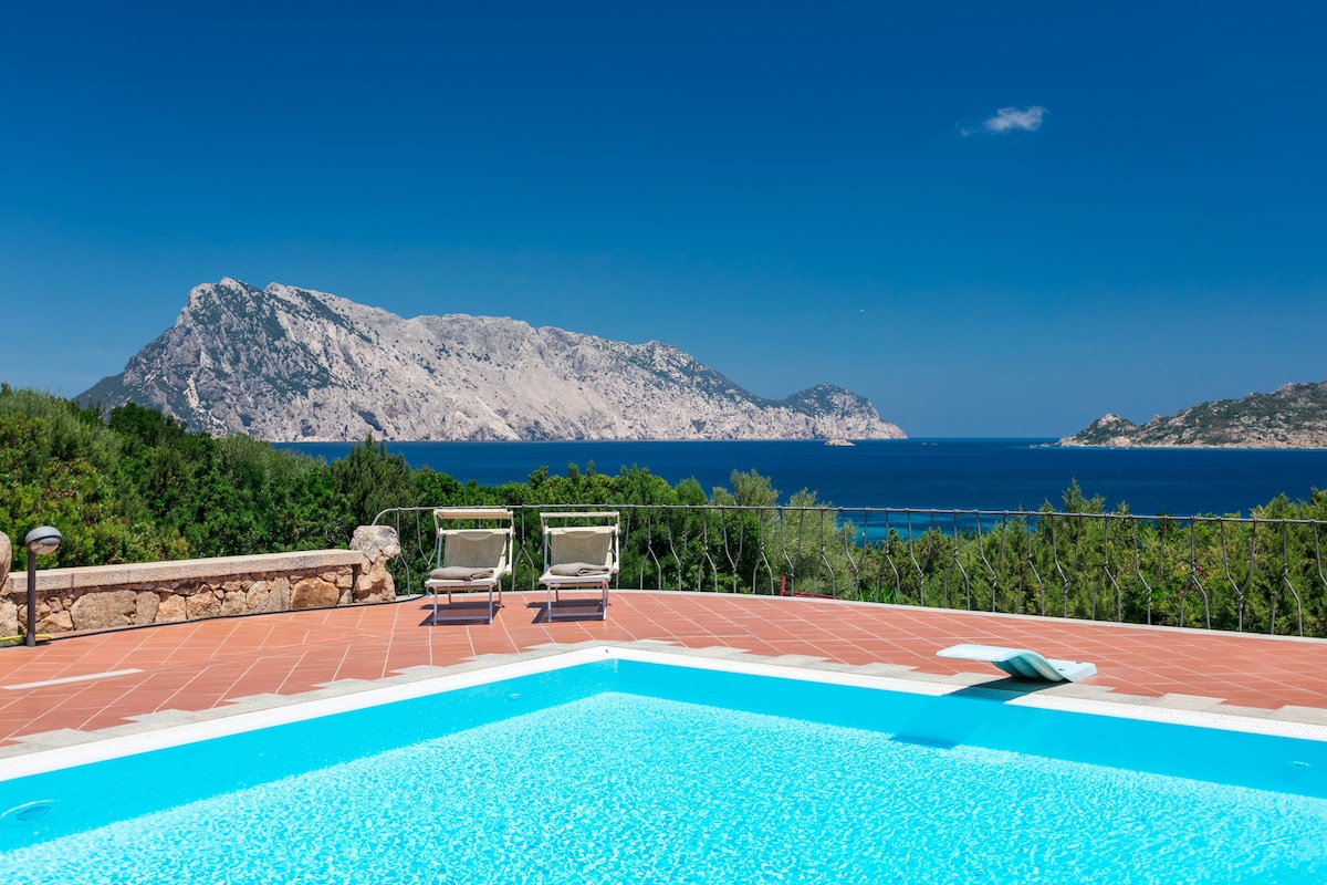 A panoramic view showcases a shimmering pool surrounded by a sun terrace. Two loungers are positioned near the edge, inviting relaxation while overlooking the coastline and the distant mountains. The clear blue sky and vibrant waters enhance the sense of tranquility in this outdoor space.