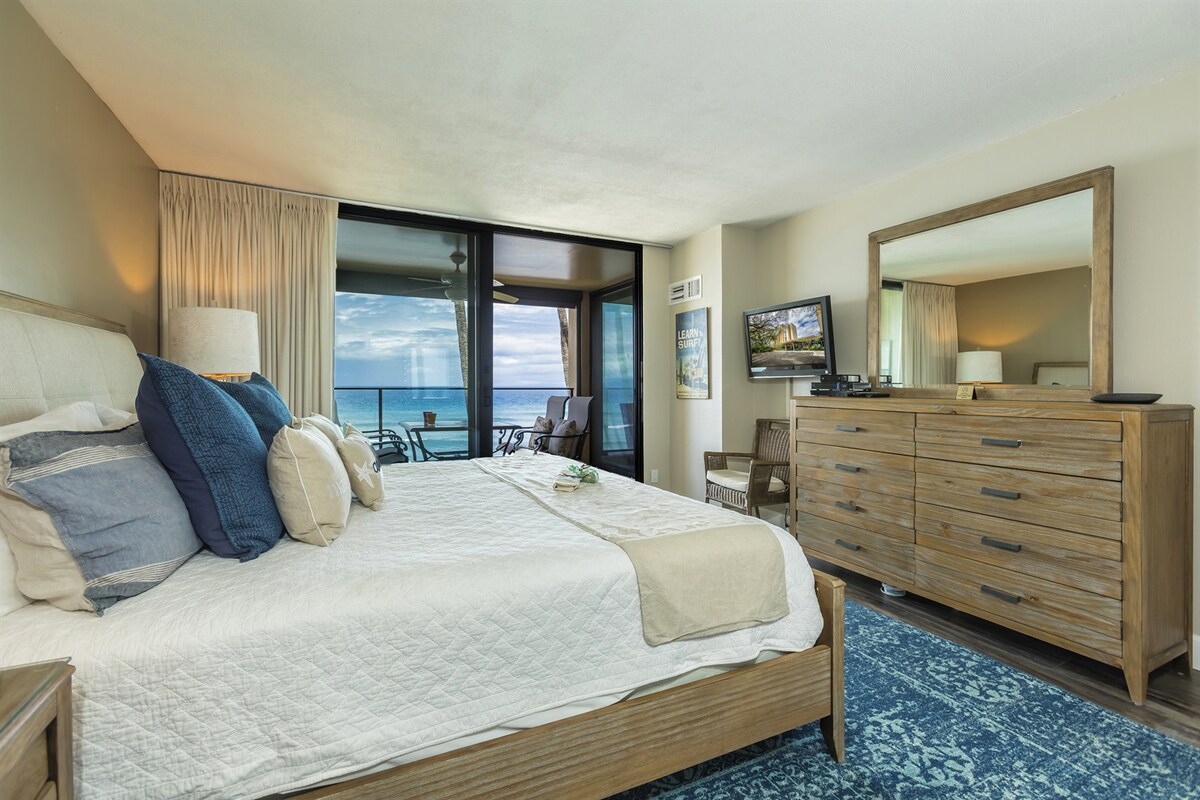 The bedroom features a spacious bed with soft linens and a selection of pillows. A large mirror reflects natural light, and a wooden dresser is positioned against the wall. Floor-to-ceiling windows offer views of the ocean, with an outdoor seating area visible on the lanai.