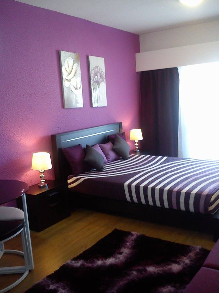 A cozy sleeping area features a double bed with striped bedding and accent pillows. The walls are painted in a rich purple hue, complemented by two floral art pieces. Soft lighting from bedside lamps creates a welcoming ambiance, while a window provides natural light.