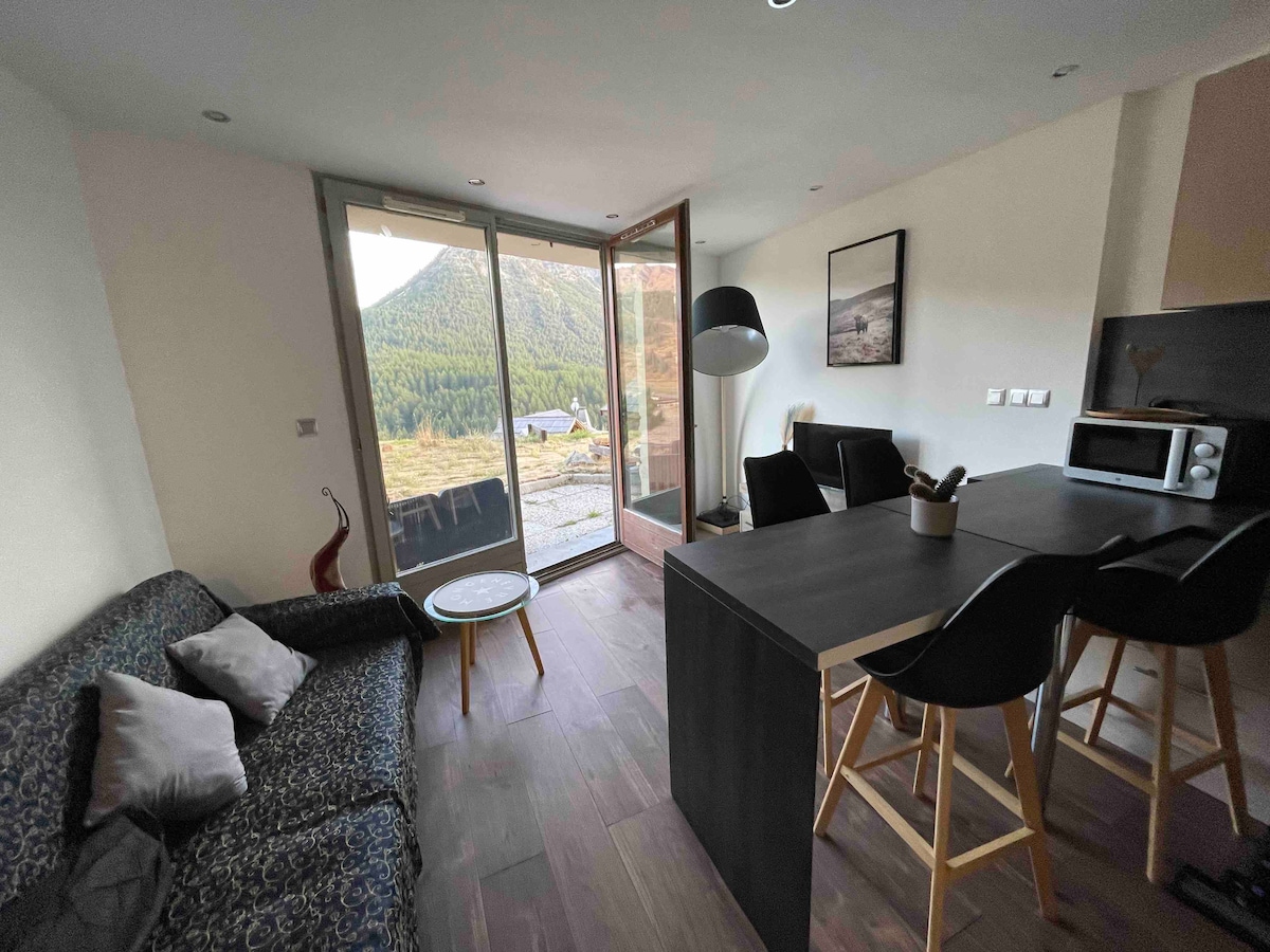 An open living space is presented with a modern kitchen featuring black chairs at a wooden table. A sofa with decorative pillows is visible, and large glass doors provide an unobstructed view of the outdoor scenery. Natural light fills the area.