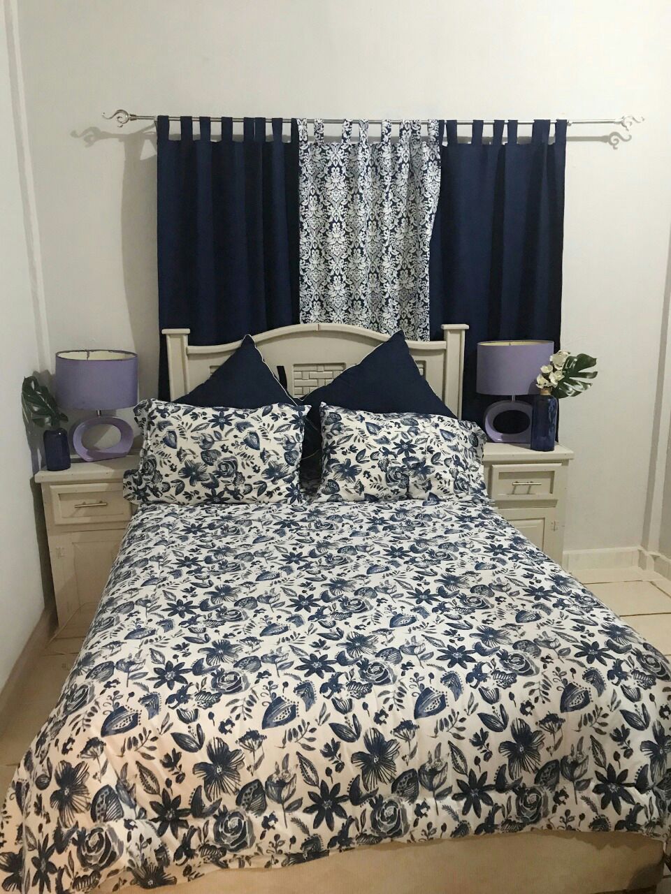 A bedroom is presented with a spacious bed covered in a floral blue and white bedspread, featuring two decorative pillows. Matching nightstands are positioned on either side, each topped with purple lamps. Dark curtains frame a patterned window treatment above the bed.