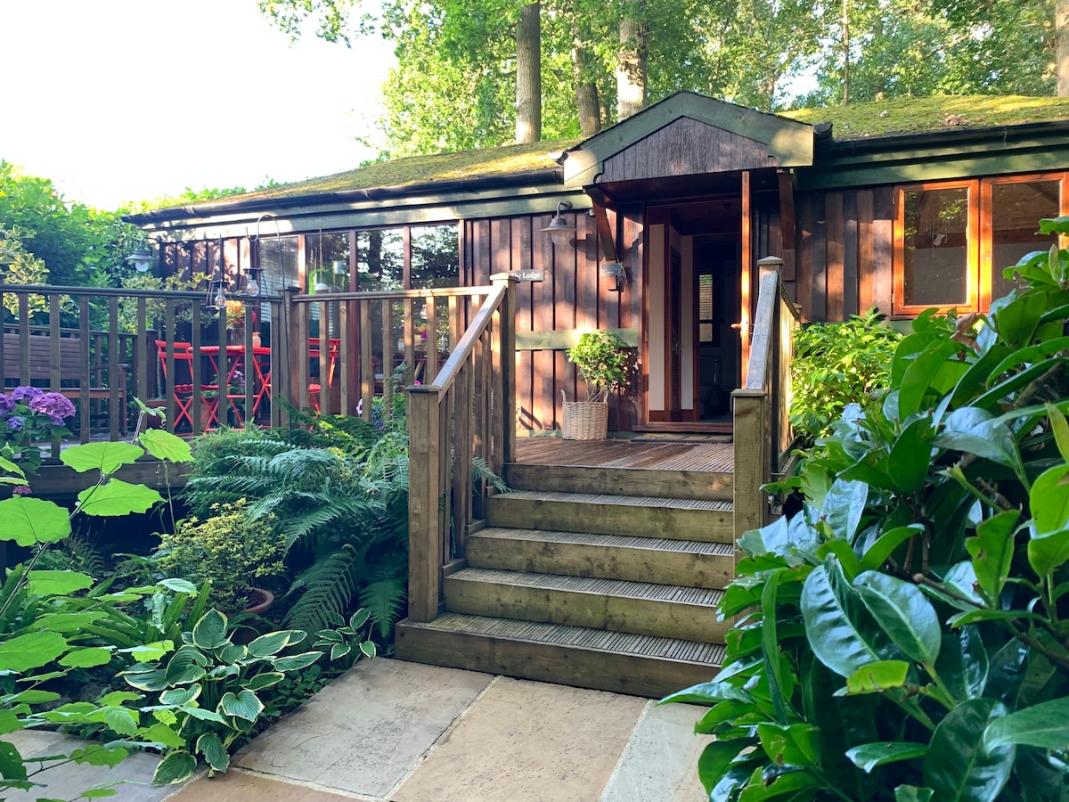 The entrance to the wooden lodge is surrounded by lush greenery and plants. A set of steps leads up to a welcoming doorway, framed by large windows that overlook the garden. A small table and chairs can be seen on the patio area to the side.