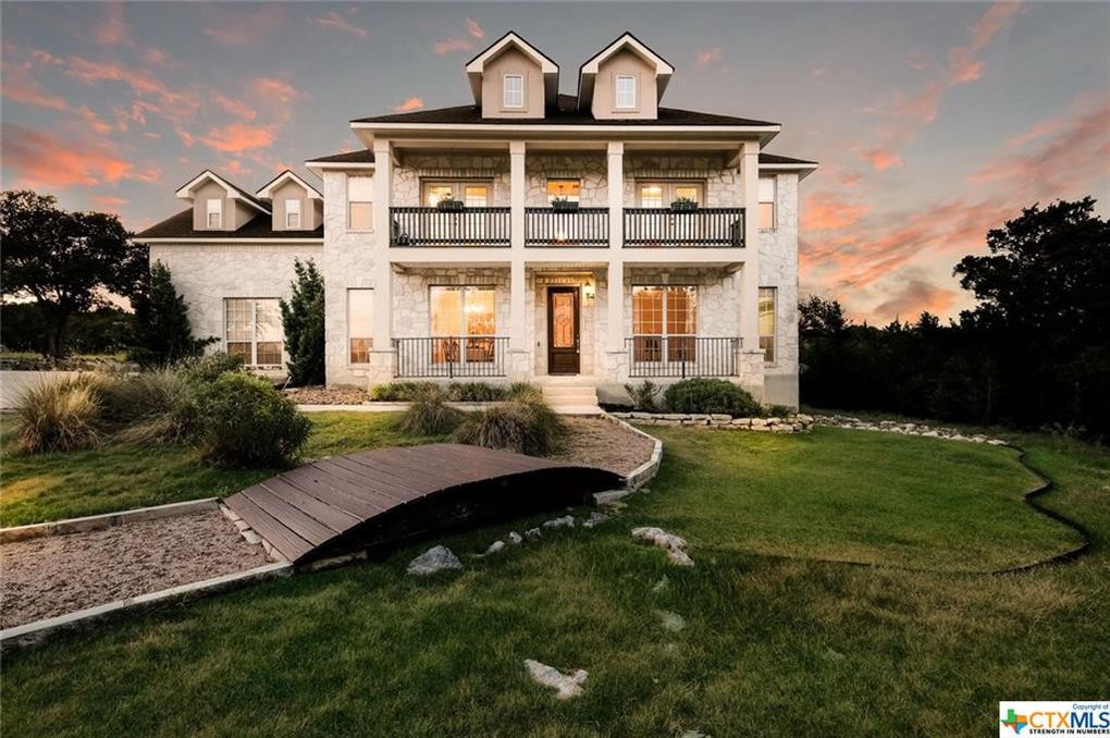 A spacious two-story home is featured, showcasing a blend of natural stone and light-colored siding. The front yard is adorned with gentle landscaping and a wooden walkway leading to the entrance. Balconies on both levels provide outdoor space, framed by a sunset-lit sky.