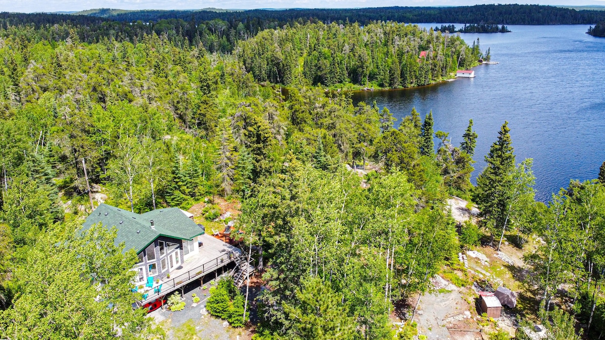 An aerial view showcases a spacious cabin surrounded by lush greenery, situated near a serene lake. The expansive deck and nearby outdoor seating areas are visible, emphasizing the natural landscape and tranquil waters.