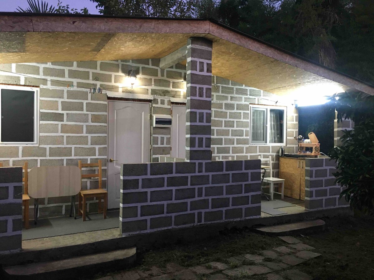 A simple, single-story structure features a covered patio area with a small dining table set for four. Walls are constructed with light-colored blocks, and large windows provide ample natural light. An outdoor kitchen space is visible, integrated with the patio layout.