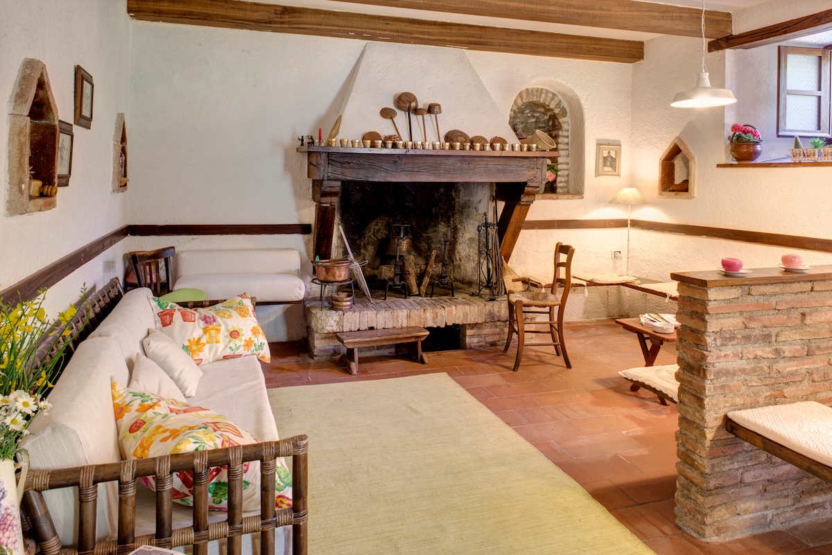 A spacious living area features a rustic fireplace as its focal point, complemented by a cozy sofa and a textured area rug. Soft lighting is provided by a hanging lamp, while wooden beams enhance the room's warm, inviting character.