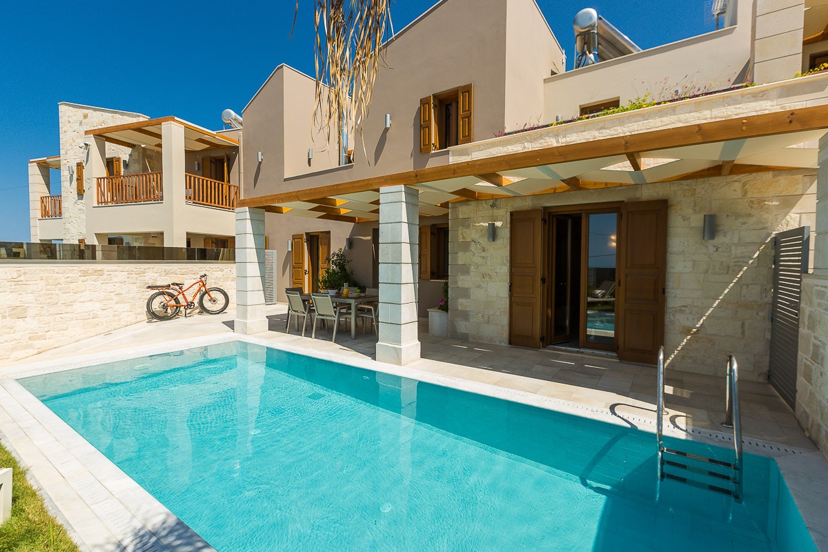A private heated pool is surrounded by a sunlit terrace featuring a small dining area and stylish seating. The villa's modern architecture is complemented by natural stone, while large doors provide access to the indoor spaces. Lush greenery adds a touch of nature to the setting.