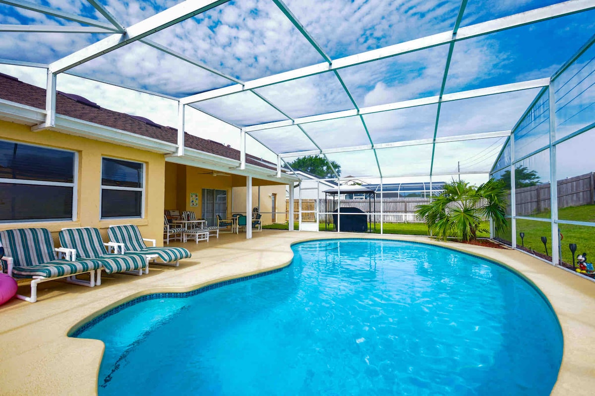 A spacious pool area is showcased under a protective screen. A large pool is bordered by a textured deck, with several lounge chairs arranged nearby. Lush greenery is visible in the background, and a covered patio area offers shaded seating.