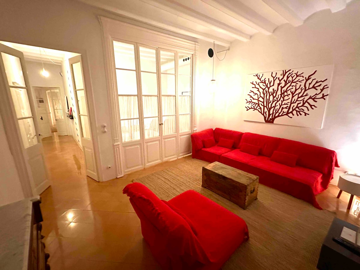 A spacious living area is furnished with vibrant red sofas, creating a bold yet comfortable seating arrangement. Natural light filters in through large glass doors, which lead to a balcony. A rustic wooden coffee table adds a touch of warmth to the room.