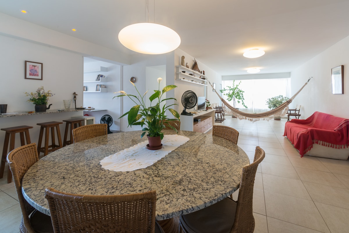 A spacious dining area features a round granite table surrounded by wicker chairs. A green plant in the center complements the light decor. In the background, a hammock is suspended for relaxation, and a cozy sofa is positioned nearby, creating an inviting atmosphere.