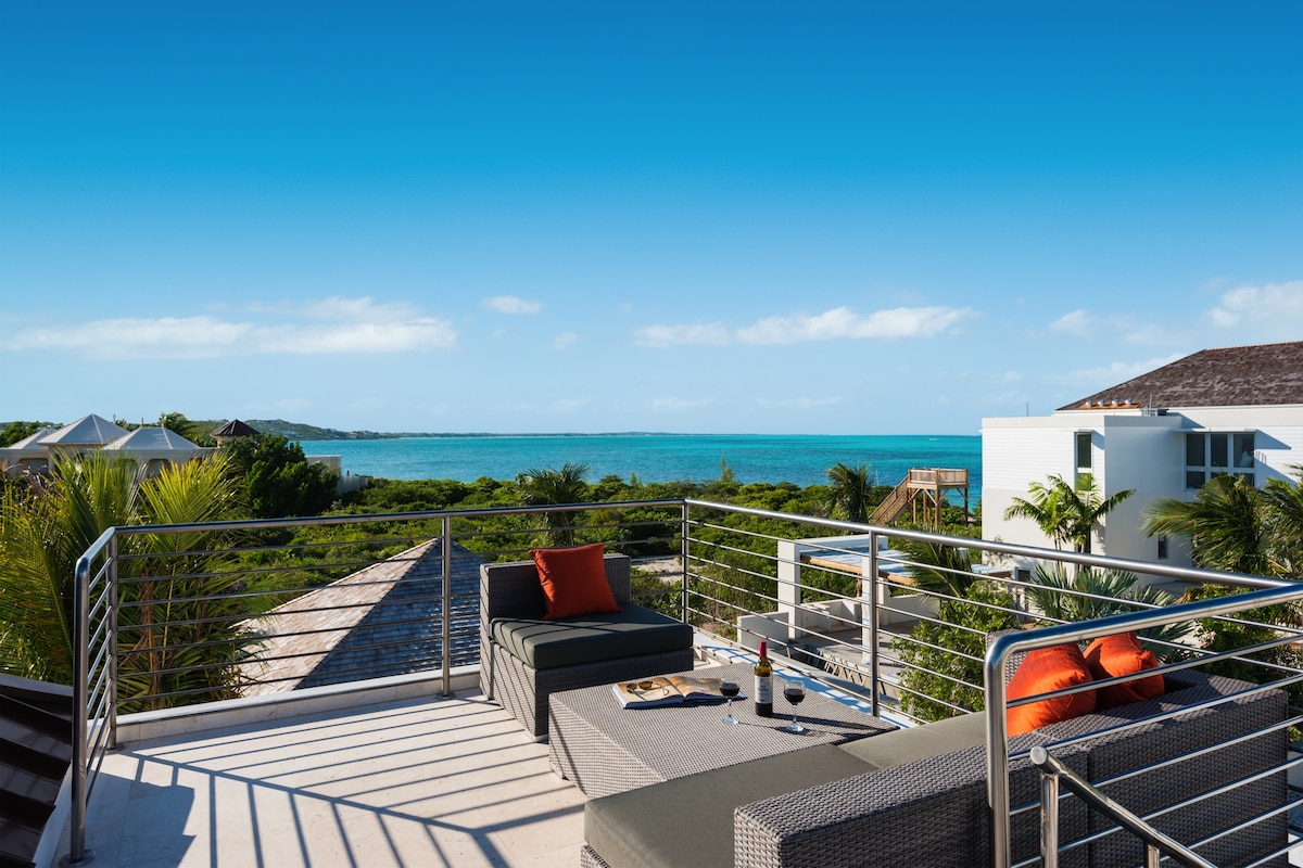 A spacious rooftop deck features comfortable seating with orange accents, overlooking a vibrant blue ocean and lush greenery. The area is designed for relaxation and outdoor dining, with a serene view of the coastal landscape under a clear sky.