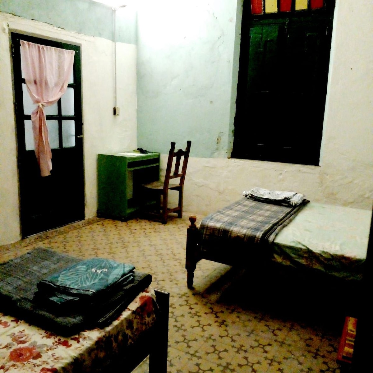 A simple room features two beds placed on opposite sides, complemented by a chair and a small green desk. Light filters through a window adorned with colorful glass panes. A pink curtain adds a soft touch to the doorway.